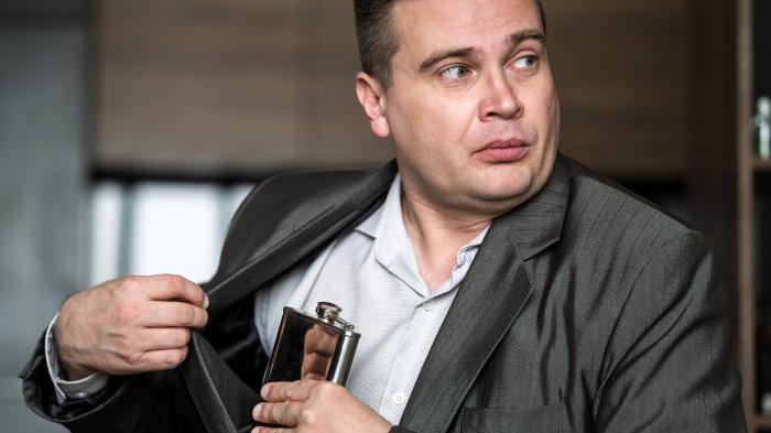 Бизнесмен выглядит встревоженным, вынимая фляжку из куртки на работе, скрывая алкогольную зависимость на рабочем месте.